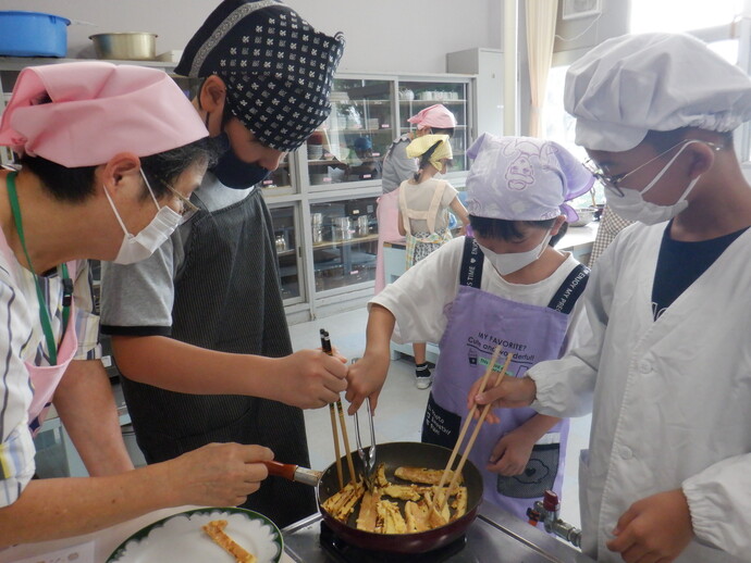 調理実習室のような場所で、エプロンと三角巾を身に着けた子どもたちと大人がフライパンを囲み、菜箸やトングを使って調理をしている様子の写真