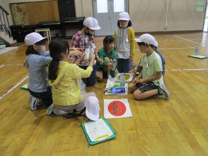 体育館の床に座った子どもたちがグループで作業をしており、中央にはペットボトルや牛乳パックなどの資材と「可燃ごみ」と書かれた赤い円の紙が置かれている様子を写した写真