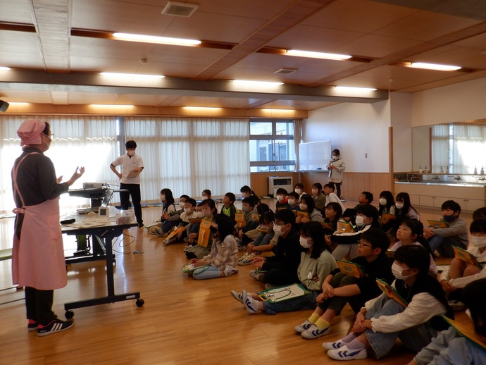 明るい多目的室のような教室で、ピンク色のエプロンと三角巾を身につけた講師の女性が、床に座りながらメモを取る多くの小学生たちに向けて、プロジェクターを活用しながら食育や調理に関する話を熱心に伝えている様子を捉えた写真