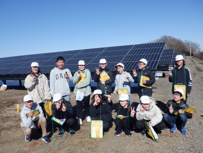 屋外の太陽光発電施設で、複数の人物がソーラーパネルの前に立ったりしゃがんだりして記念撮影をしており、全員が白い帽子や防寒着を着用し、手には黄色いバインダーや筆記用具を持っている様子が写された写真