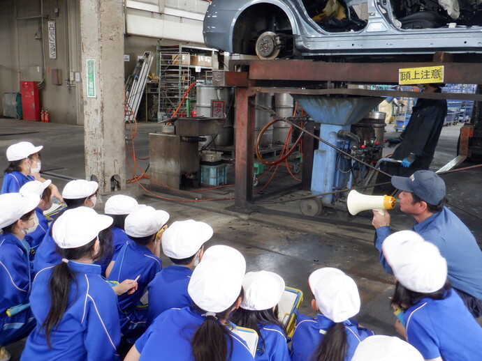 自動車整備工場のような屋内で、白い帽子と青い服を着た児童が複数座り、ノートを持って説明を聞いており、前方には車体がリフトで高く持ち上げられ、下には金属製の機械やホースが配置され、黄色い「頭上注意」の表示が見える様子の写真