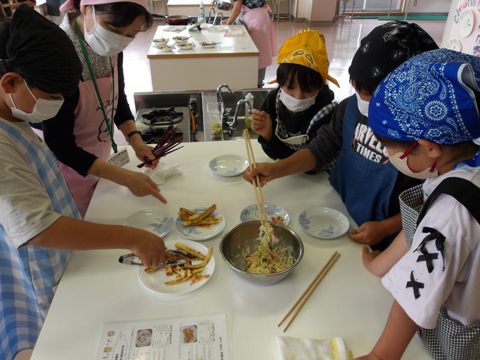 調理室でエプロンと三角巾を着けた子どもたちが、白いテーブルの上で金属ボウルに入った切り干し大根のサラダを菜箸で取り分け、隣にはパンのみみフレンチトーストを盛り付けた皿や空の小皿が並び、手前にはレシピが置かれている様子の写真