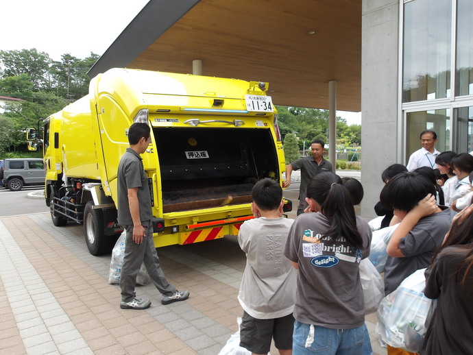 校舎の玄関前で黄色いごみ収集車の後部が開いた状態で停車し、作業服姿の職員が説明を行っており、複数の児童が袋を手にして車両の仕組みを見学している様子の写真