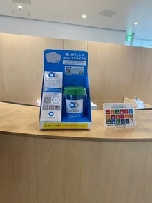 緑の蓋が付いた透明のカップが入った、青いコンタクトレンズ空ケースの回収ボックスが、SDGsのロゴが並んだ小さなパネルと並んで、市民交流センターtette総合案内の木製カウンターの上に置かれている写真