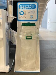 階段下のスペースに透明の蓋つきごみ箱が入った白いボックスが置かれ、上に回収ボックスの案内が設置された、コンタクトレンズ空ケースの回収ボックスの写真