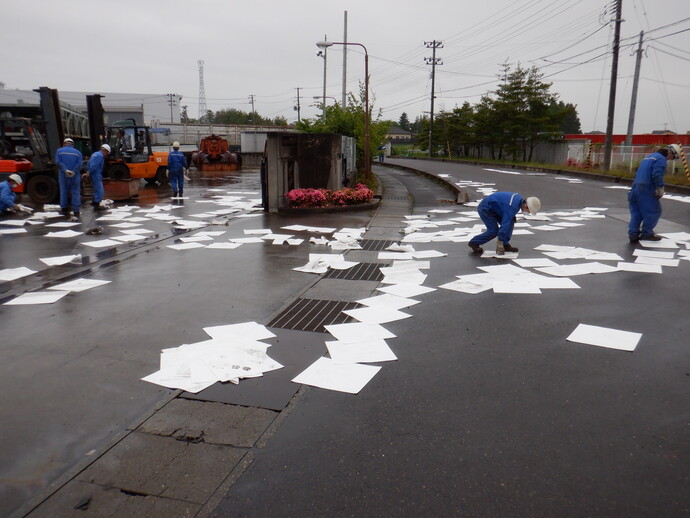 雨の工場敷地で多数の油吸着マットが濡れた地面一面に散乱し、作業員が広い範囲を歩き回って設置している様子の写真