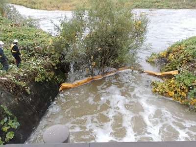 増水した川で流木防止用のオイルフェンスが木に固定され、作業員が護岸で状況を確認している様子の写真