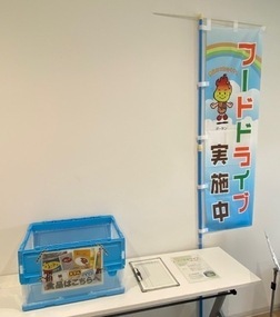 白い壁際に設置された受付デスクの上に、青い食品回収ボックスと受付名簿が置かれ、その横には虹とキャラクターが描かれた「フードドライブ実施中」という青い幟が立てられている、屋内の特設コーナーの様子の写真