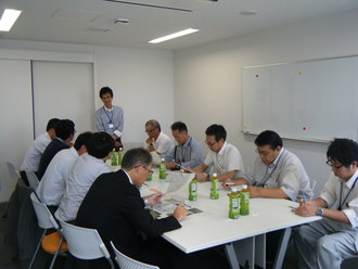 会議室にて、複数の関係者が長テーブルを囲んで座り、手元の資料を読み込んだりメモを取ったりしながら、一人の男性による報告や説明に耳を傾けている、真剣な雰囲気の会議風景を捉えた写真