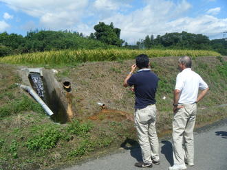 黄金色の稲穂が広がる田んぼの土手にて、二人の男性がコンクリート製の排水路から勢いよく水が流れ落ちているのを観察しながら話をしている様子の写真