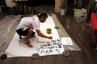 室内で長渕剛さんが大きな紙の上にしゃがんで筆を運び、周囲に道具を配置して書を仕上げている様子の写真