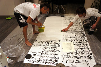 広い床に養生シートを敷いた室内で二人が大きな紙に向かい、筆や重りを使って共同で書を仕上げている様子の写真