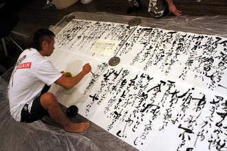室内で長渕剛さんが床に広げた大きな紙へ重りを添えて筆を運び、力強い書を仕上げている様子の写真