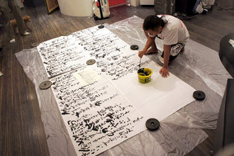 室内で床一面に広げた大きな紙を重りで固定し、長渕剛さんが筆と墨を使って力強い筆致で書を仕上げている様子を捉えた写真