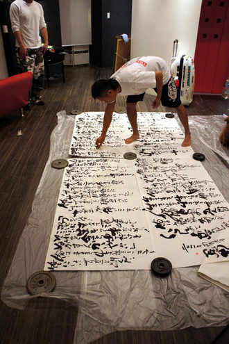 室内で床一面に広げた大きな書道作品を重りで固定し、長渕剛さんが書の上に足を乗せ、中腰になって筆を使って文字を書き進めている様子の写真