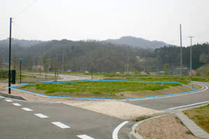 広い敷地のロータリー状に舗装された道路に面した場所に、青い線で楕円形に囲まれた緑の草地があり、その奥には山と木々が見える区画 No.12の写真