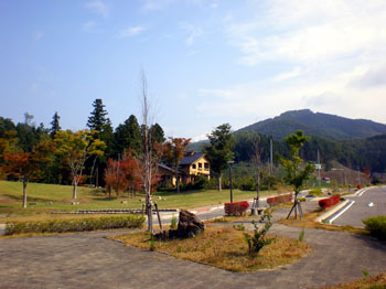 黄色や赤の紅葉を背景に、広大な芝生とその奥にログハウス風の建物が建ち、右手前には舗装された道路と散策路が伸びている秋のガーデンタウン虹の台の写真