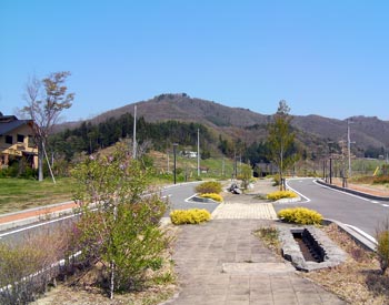 青空の下、舗装された歩道と道路が曲線を描いて伸びており、その先の背景には緑の木々が生い茂る低山が広がり、道の脇には黄色い花をつけた低木などが植えられている春のガーデンタウン虹の台の写真