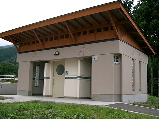 ベージュの外壁と木製の屋根が特徴的な建物で、中央には多目的トイレのドアがあり、左右に入口が分かれた屋外トイレの外観写真