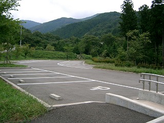 青々とした山々を背景に、アスファルト舗装の駐車場があり、手前には車椅子利用者のための優先駐車スペースやスロープが設置されている写真
