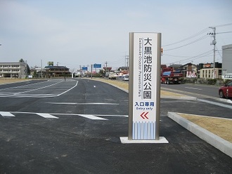「大黒池防災公園 入口専用」と書かれた白い縦長看板が手前に立ち、その奥に新しく舗装されたアスファルトの道路と広い駐車場が広がる公園入り口を映した写真