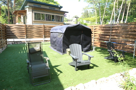 木製の柵に囲まれた人工芝のスペースに、黒い大型のテントサウナが設置され、その手前にはリクライニングチェアやプラスチック製の椅子が並べられており、奥には木造の建物が建っている屋外のサウナ施設を捉えた写真