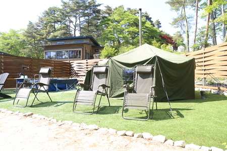 抜けるような青空と新緑の木々に囲まれた人工芝の広場に、煙突の付いたカーキ色の大きなテントサウナが設置され、その手前にはゆったりとくつろげる3台のリクライニングチェアが並んでいる開放的な屋外サウナ施設を捉えた写真