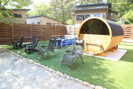 木製の柵で囲まれた芝生エリアに、丸い樽型が特徴的なバレルサウナが設置され、その手前にはゆったりとしたリクライニングチェアや水風呂用のプールが並び、背景には木立の中にモダンな建物が点在している屋外サウナ施設を捉えた写真