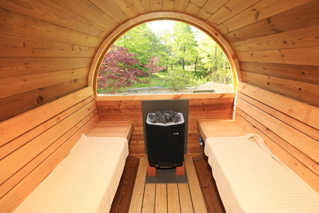 木の温もりに包まれた円形のバレルサウナ内部で、中央にはサウナストーンが積まれた黒いヒーターが鎮座し、左右のベンチには白いマットが敷かれ、正面の大きな半円形の窓からは新緑の美しい景色を望むことができる室内写真