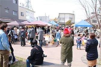 晴天に恵まれた広々とした屋外広場で、青や茶色のパラソルが並ぶ露店を多くの人々が訪れているイベント風景の写真