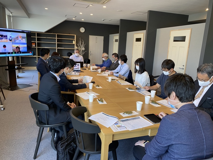 木製の大きな会議テーブルを囲んでマスク姿の参加者が着席し、資料と飲み物を前に会議を行い、左側の大型ディスプレイにオンライン参加者が表示されている会議の写真