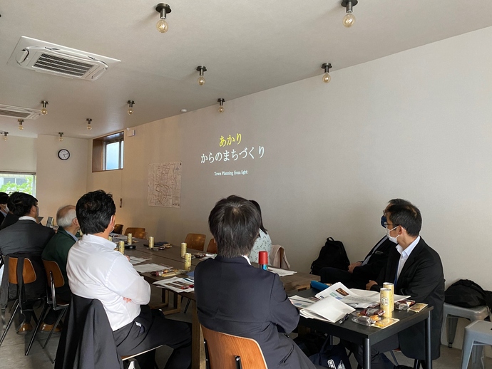 白い壁に「あかりからのまちづくりと」文字が映し出され、木製テーブルに座った参加者たちがマスクを着用し資料を見ながら会議をしている写真