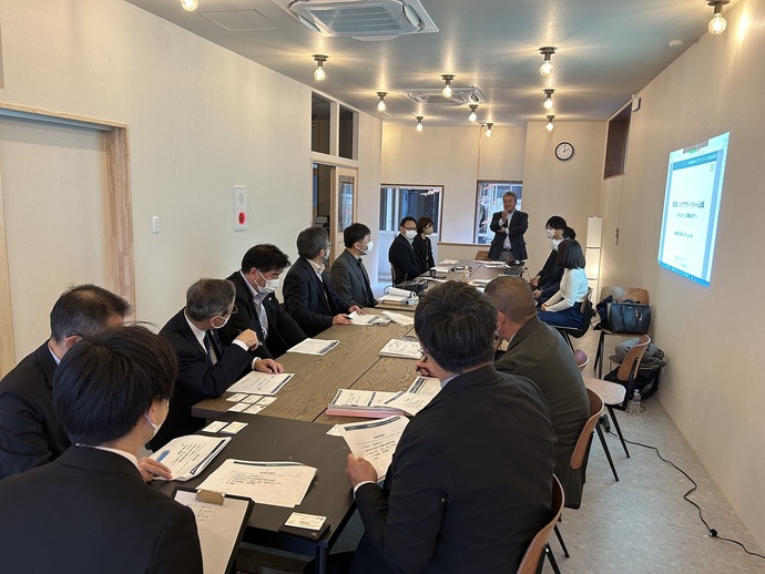 白い壁の会議室で、マスク着用のスーツ姿の参加者たちが大きな木製テーブルを囲んで資料を見ながら会議をし、右側の壁にスクリーンが投影されている会議の写真