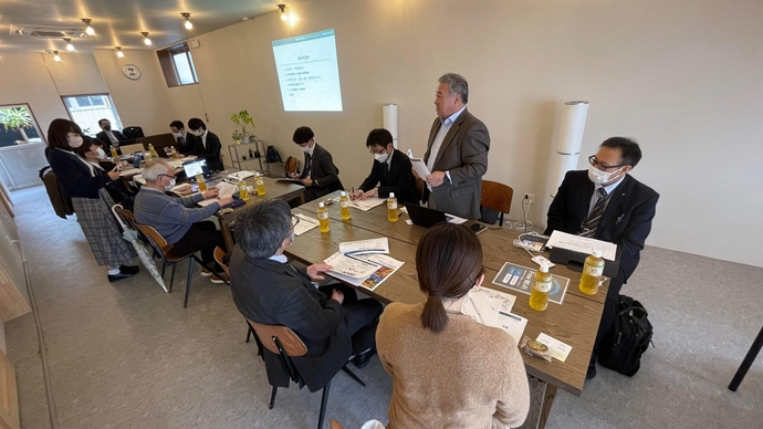 白い壁の明るい会議室で、木製の大きなテーブルを囲んでマスク着用の参加者が着席し、立っているスーツ姿の男性が説明をし、壁のスクリーンに資料が投影されている写真