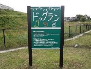 緑色の背景に白文字で大きく「ドッグラン」と書かれ、可愛らしい犬のイラストや利用上の注意が細かく記された案内看板が、フェンスに囲まれた芝生広場の手前に設置されている様子を正面から捉えた写真