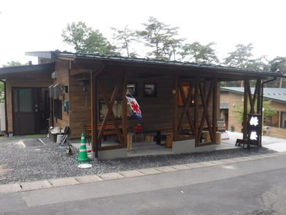 緑の木々を背景に建つ木造平屋建てで、屋外テラス席に氷と書かれた旗がかかった、飲食施設の外観写真