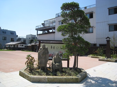 建物前のタイル敷きの広場の一角に、松の木と共に二体の男性の石像が設置されており、背後に「結の辻」と書かれた看板が立っている写真