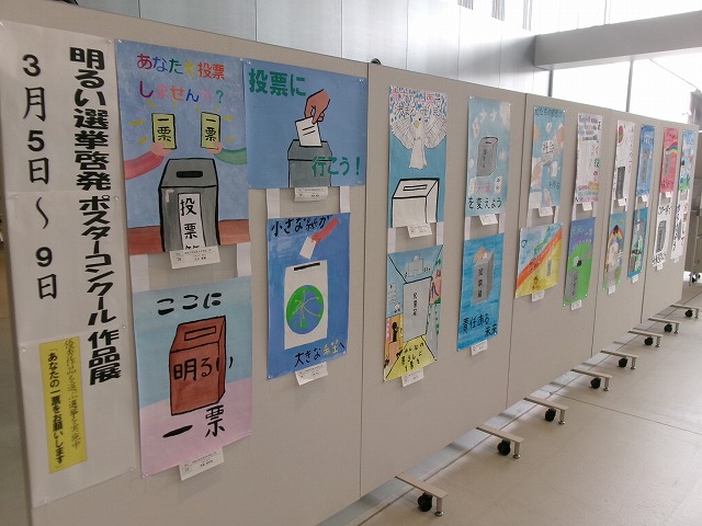 複数並んだ展示パネルにたくさんのポスターが展示され、左端に「明るい選挙啓発ポスターコンクール作品展」と書かれた展示の様子の写真