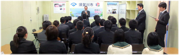 「須賀川市」のロゴが入ったパネルを背にマイクを持つ男性と、その前方で椅子に座り話を聞くリクルートスーツ姿の男女や制服を着た学生たちの後ろ姿を捉えた採用内定者説明会の様子の写真