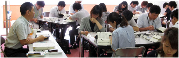 大勢の男女が複数のテーブルに分かれて座り、手元の資料や書籍を真剣な表情で読み込んだりメモを取ったりしながら、集中した様子でグループワークに取り組んでいる、地方自治法、地方公務員法研修の様子の写真