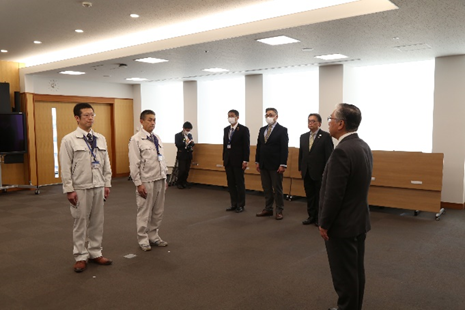 広々とした会議室で、作業服を着た2名とスーツ姿の男性が向き合っており、そばでスーツ姿の複数名の関係者が見守っている様子の写真