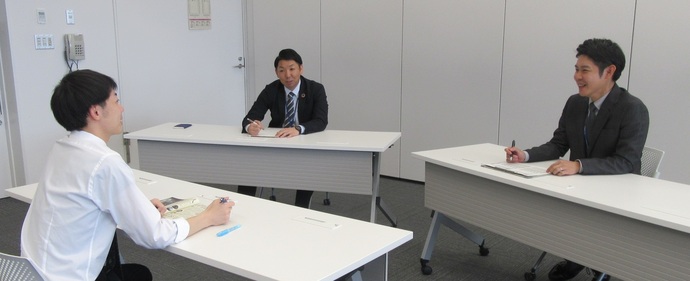 清潔感のある会議室で、白シャツ姿の男性が手前に座り、対面に座るスーツ姿の二人の男性と、資料やペンを手に持ちながら穏やかな表情で面談や打ち合わせを行っている様子の写真