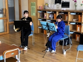 掲示物が並ぶ明るい教室内で、黒いTシャツや青いジャージを着用した2人の生徒が、椅子に座って譜面台の楽譜を真剣に見つめながらホルンを構え、パート練習や音出しに集中して取り組んでいる風景の写真