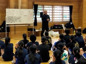 体育館で黒いジャージ姿の指導者がホワイトボードの前に立ち、床に座って並んだ黒い練習着姿の多くの参加者たちに向けて説明しているミーティング風景の写真