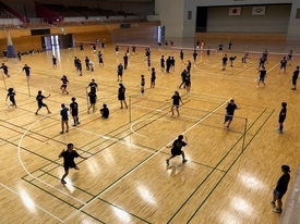 広い体育館で、黒いTシャツを着た多数の参加者が複数のコートに分かれてバドミントンの練習に励んでいる様子を、アリーナ全体が見渡せる高い位置から撮影した写真