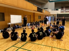 体育館の床に座った金色の背番号入り黒Tシャツ姿の参加者たちが、ホワイトボードの前に立つ指導者の説明を真剣な様子で聞いているミーティング風景の写真