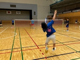 体育館のバドミントンコートで、背中にピンクのロゴが入った青いTシャツを着た選手が大きくジャンプして、ネット越しの相手に向かってスマッシュを打とうとしている瞬間の写真