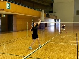 体育館のバドミントンコートで黒いTシャツを着た選手が頭上でラケットを構えてシャトルを打つ動作をしており、ネット越しの白いシャツの選手がそれに応じようと構えている練習風景の写真