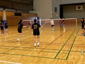 体育館のコートで金色の背番号が入った黒いTシャツのペアが、ネット越しの相手選手とダブルスの試合形式で練習をしている様子の写真