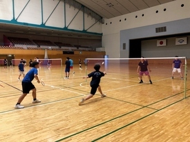 体育館のバドミントンコートで青いTシャツと黒いTシャツを着たペアがネット越しの相手とダブルスの試合形式で対戦しており、シャトルを打ち返すために構えている練習風景の写真